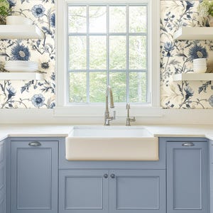 May include: A white farmhouse kitchen with blue cabinets and a white porcelain farmhouse sink. The walls are covered in a floral wallpaper with blue and white flowers.