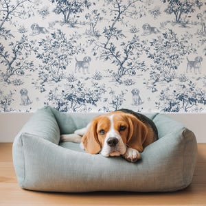 Puede incluir: Un perro beagle descansa en una cama para mascotas azul claro. El perro tiene marcas marrones, negras y blancas. El fondo presenta un papel pintado toile de Jouy azul y blanco con patrones de perros y flores. El suelo es de madera.