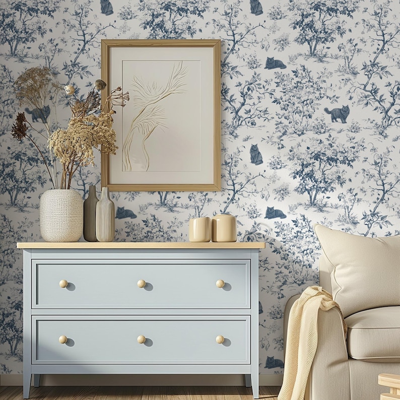 May include: A pale blue chest of drawers with two drawers and wooden knobs. It has a light wood top with a vase of dried flowers, two bottles, and two small cream-coloured ceramic cups. The background is blue and white wallpaper with a cat and tree design.