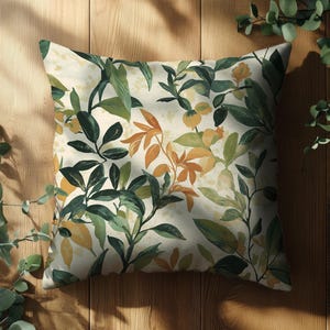 May include: Square decorative pillow with a botanical print. The pillow features a cream-colored background with a pattern of green, yellow, and orange leaves and branches. The pillow is placed on a wooden surface, with leafy branches in the background.