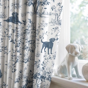 Op de afbeelding: Witte gordijn met een blauw toile de Jouy-patroon met honden en bloemmotieven. Een wit keramieken hondenbeeld en een vaas met witte bloemen staan op een vensterbank. Het thema is hondenthema woondecoratie.