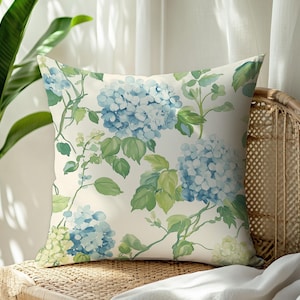Puede incluir: Una almohada blanca con un patrón floral con hortensias azules y verdes. La almohada está sentada en una silla de mimbre.