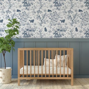 May include: A wooden cot with vertical bars, a mattress, and pillows. The cot is in front of a blue wall with a white and blue cat and tree pattern wallpaper. A potted plant is to the left.
