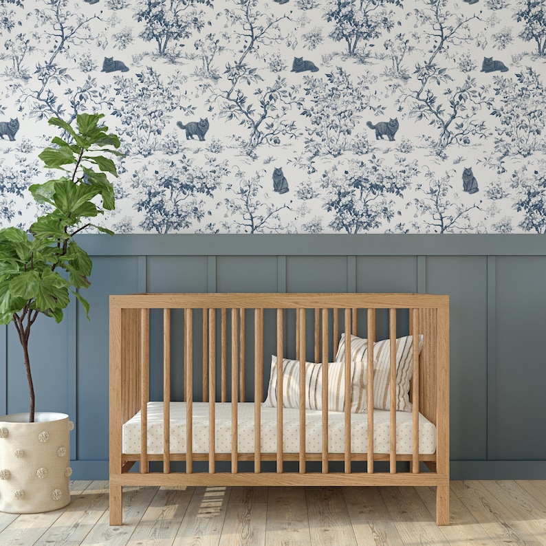 May include: A wooden cot with vertical bars, a mattress, and pillows. The cot is in front of a blue wall with a white and blue cat and tree pattern wallpaper. A potted plant is to the left.