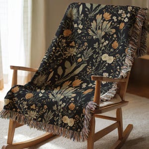 Puede incluir: Una manta de tejido negro con un patrón floral que presenta varios tonos de marrón, amarillo y verde. La manta está drapeada sobre una mecedora de madera.