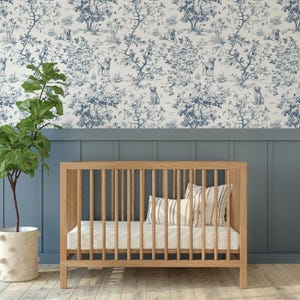 May include: A wooden crib with vertical slats, a mattress, and pillows. The crib is set against a blue wall and a wallpaper with a blue and white toile pattern featuring dogs and trees. A potted plant is on the left.
