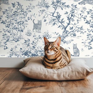 Könnte beinhalten: Eine braune Tabbykatze ruht auf einem beigen Kissen. Die Katze befindet sich vor einer Tapete mit einem blau-weißen Blumen- und Katzenmuster. Der Boden ist aus Holz.
