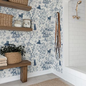 May include: A bathroom scene featuring a wooden bench with folded towels and a woven basket. The wall is adorned with wallpaper depicting blue dogs in a pastoral scene. A leather leash hangs on the wall, and a shower with gold fixtures is visible.