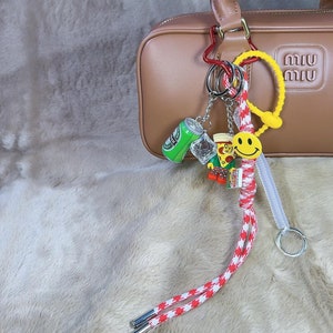 May include: A brown leather handbag with a gold metal handle and a gold metal zipper. The bag has a charm attached to it with a smiley face, a miniature can of soda, a miniature pizza, and a miniature Lego figure. The charm is attached to the bag with a red and white braided cord.
