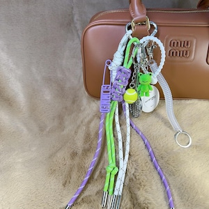 Op de afbeelding: Een bruine leren handtas met een verscheidenheid aan kleurrijke sleutelhangers aan de handgreep. De sleutelhangers omvatten een groene kikker, een tennisbal, een wit en groen touw, een paars touw en een zilveren veiligheidsspeld. De tas heeft een gouden rits en een gouden gesp.