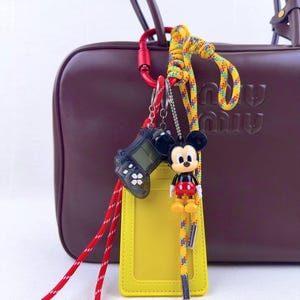 May include: A brown handbag with a red carabiner clip holding a yellow lanyard, a black handheld game, and a Mickey Mouse figurine. The lanyard has a yellow ID holder and a colourful rope. The bag has a subtle logo.