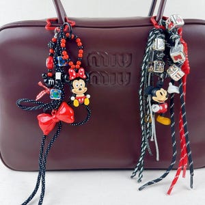 May include: Two bag charms with Mickey and Minnie Mouse figures. One has a red bow, beads, and a Minnie Mouse figure. The other has Mickey Mouse and cube-shaped charms. Both have black and white cords, hanging in front of a brown handbag.