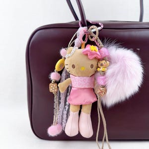 May include: A Hello Kitty plush keyring in a pink dress and boots, decorated with a pink flower, beads, and a fluffy pink pom-pom. The keyring is attached to a burgundy handbag.