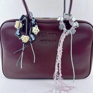 May include: A burgundy leather handbag with a top handle and the embossed brand logo. The bag is accessorized with two decorative charms, one with heart and star shapes, and the other with silver cubes and pink crochet.