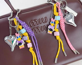 May include: A brown leather handbag with the brand name embossed on the front. Attached are several keychains with cartoon duck figures, silver heart charms, and colorful braided cords in yellow, purple, and pink.