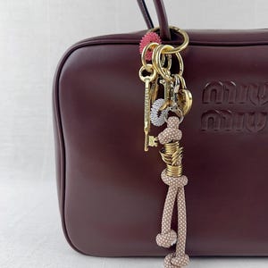 Peut inclure: Un sac à main en cuir bordeaux avec des accessoires dorés et un porte-clés décoratif. Le porte-clés comprend une clé dorée avec le texte "FIRST FLOOR", un charme en forme de cœur et une corde à pois beige et blanche.