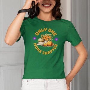 Op de afbeelding: Een groen t-shirt met korte mouwen en een grafische print van gestapelde boeken, oranje pompoenen en gele bloemen. De tekst "ONLY ONE MORE CHAPTER" is in het geel rond de centrale illustratie gedrukt, geaccentueerd door twee kleine paarse bloemen.