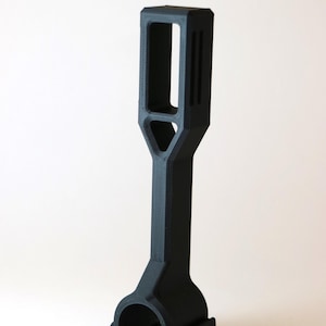 Pode incluir: Uma montagem de mira telescópica de plástico preto com uma abertura retangular e um recorte triangular. A montagem tem um anel na parte inferior para fixar a uma mira telescópica.