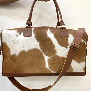 May include: A brown and white cowhide leather duffel bag with a brown leather shoulder strap and handles. The bag has a rectangular shape with a zipper closure and metal hardware. The bag is held by a person's hand.