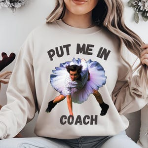 May include: A beige sweatshirt with the text "PUT ME IN COACH" printed in black. A photo of a man in a tutu is printed on the sweatshirt.