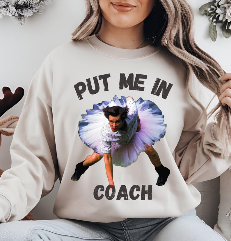 May include: A beige sweatshirt with the text "PUT ME IN COACH" printed in black. A photo of a man in a tutu is printed on the sweatshirt.