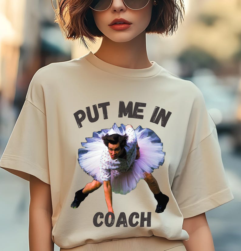 May include: A beige t-shirt with the text "PUT ME IN COACH" printed on the front. The text is in black and there is a picture of a man in a tutu with the text "PUT ME IN COACH" printed on the front.