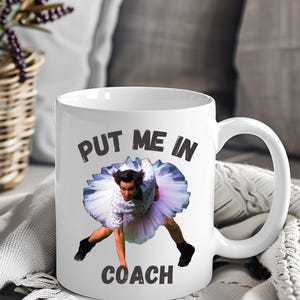 May include: White ceramic coffee mug with a black and white image of a person in a tutu and the text "Put me in coach".