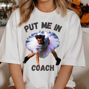 May include: White t-shirt with a graphic of a man in a tutu and the text "Put me in coach".
