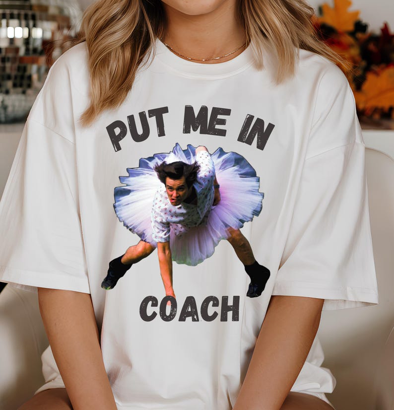 May include: White t-shirt with a graphic of a man in a tutu and the text "Put me in coach".