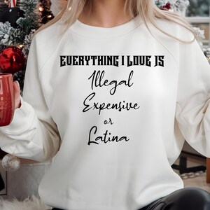 May include: White sweatshirt with black text that reads "Everything I love is Illegal Expensive or Latina".