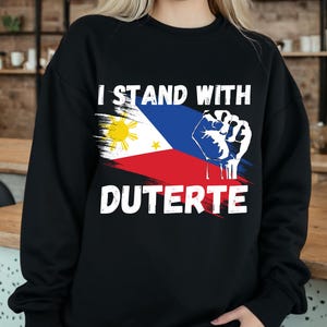 Puede incluir: Sudadera negra con un estampado gráfico blanco de la bandera filipina y un puño. El texto "I Stand With Duterte" está impreso debajo de la bandera.