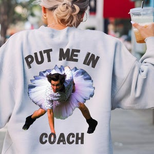 May include: A light gray sweatshirt with the text "PUT ME IN COACH" printed on the back. The text is in black and the image features a man in a tutu.