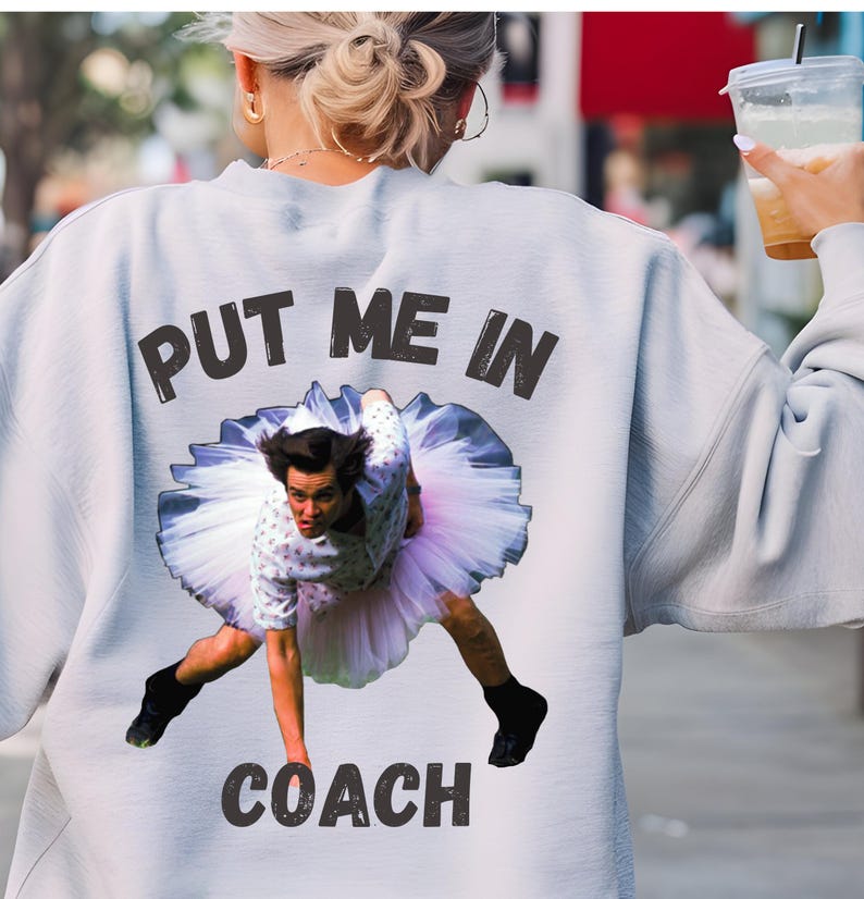 May include: A light gray sweatshirt with the text "PUT ME IN COACH" printed on the back. The text is in black and the image features a man in a tutu.