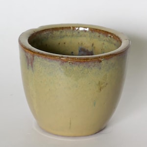 May include: A small, ceramic cup with a rounded shape. The cup has a light yellow base with a gradient of green and brown around the rim. The interior is dark. The cup is likely handmade.