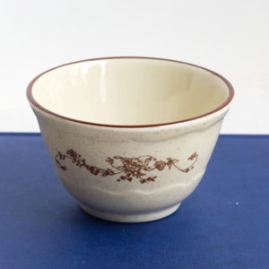 Puede incluir: Un pequeño cuenco de cerámica blanca con un borde marrón y un diseño floral se encuentra sobre un libro de tapa dura azul.