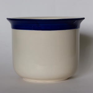 May include: A white ceramic container with a rounded base and a dark blue band around the top. The container has a simple, minimalist design, suitable for various uses.