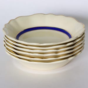 May include: A stack of six cream-coloured ceramic bowls with scalloped edges. Each bowl has a gold rim and a blue inner ring. The bowls are stacked, displaying their elegant design.