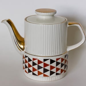 Może przedstawiać: Biały ceramiczny imbryk ze złotymi akcentami na dziobku i uchwycie. Korpus ma pionowe żłobienia, a podstawa geometryczny wzór brązowych, czarnych i pomarańczowych trójkątów. Pokrywka ma złoty uchwyt.