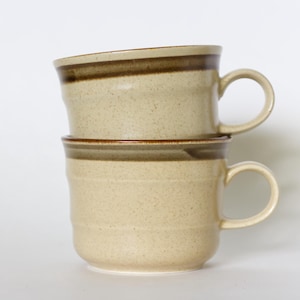 Può includere: Due tazze impilate in ceramica beige con una texture maculata. Ogni tazza ha un manico arrotondato e una fascia marrone scuro vicino al bordo. Le tazze sono su uno sfondo bianco.