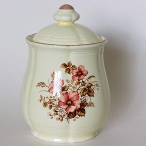 Puede incluir: Un tarro de cerámica de color crema con tapa, con un diseño floral. El tarro tiene forma redondeada con borde festoneado y un estampado floral marrón y rosa. La tapa tiene un pequeño pomo redondeado.