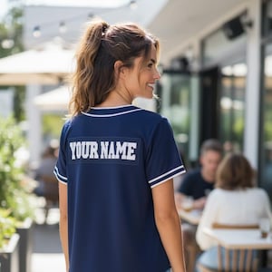 May include: Navy blue baseball jersey with white trim and the text "YOUR NAME" across the back. The jersey is paired with blue jeans. The image is taken outdoors, with a blurred background of a cafe.