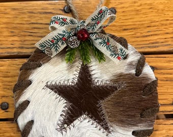 May include: Round Christmas ornament with a cowhide design. The ornament features a brown star stitched onto a white and brown hide. A decorative bow with pine needles and a red berry adorns the top, with a jute loop for hanging.