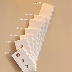 Peut inclure: Une pile de carreaux décoratifs blancs, carrés, avec des bords festonnés et des découpes circulaires. Chaque carreau est étiqueté avec sa hauteur en cm, de 6 cm à 20 cm. Les carreaux sont disposés en éventail sur un fond beige.