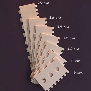 Pode incluir: Uma pilha de objetos retangulares bege, entalhados, de tamanhos variados, medindo de 6 cm a 20 cm de comprimento. Os objetos têm uma série de entalhes ao longo de uma borda e três orifícios circulares no menor. Em fundo roxo escuro.