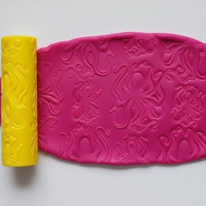 May include: A yellow rolling pin with a swirling line design sits beside a sheet of pink clay, patterned with octopuses. The clay is oval and shows detailed texture. The rolling pin and clay are for crafts.