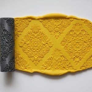 May include: A grey patterned rolling pin next to a sheet of yellow clay with an embossed floral design. The rolling pin has a repeating pattern of flowers, hearts, and other decorative elements. The clay has a similar embossed design.