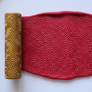 May include: A gold-colored patterned rolling pin next to a sheet of red clay with a matching pattern. The rolling pin and clay are on a white surface. The pattern resembles a brain or coral.
