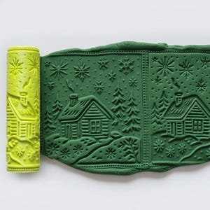 May include: A lime green rolling pin and a green clay sheet featuring a winter cabin design. The design includes cabins, trees, and snowflakes. The rolling pin has the same cabin design.