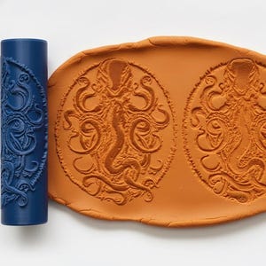 May include: A dark blue rolling pin with an octopus design next to a terracotta-coloured clay impression of the same octopus design. The octopus design is detailed and intricate, with swirling tentacles.