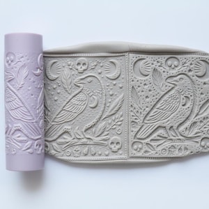 May include: A lilac clay roller and a grey clay slab, both embossed with raven and skull designs. The roller has a repeating pattern of ravens, leaves, and skulls. The slab shows two raven images with celestial and botanical elements.